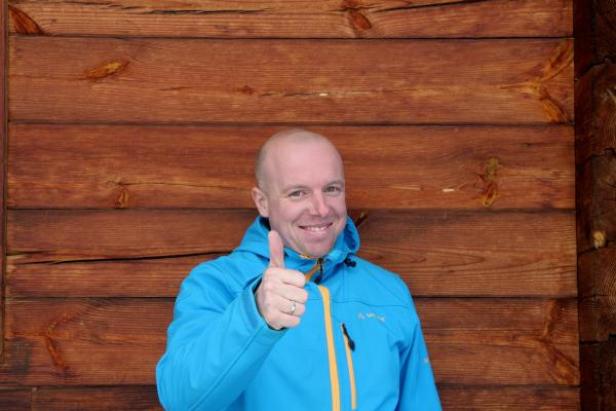 Ein Mann im blauen Anorak zeigt den Daumen nach oben vor einer Holzwand.