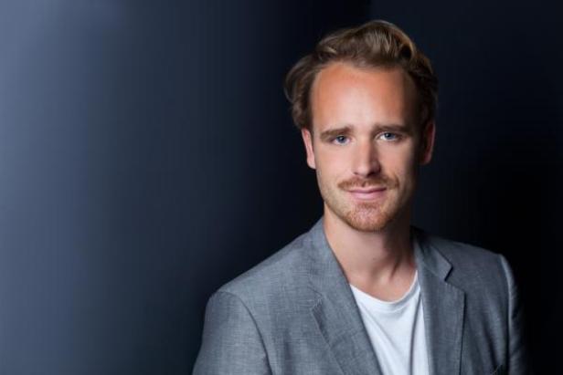 Ein Mann mit blonden Haaren und Bart trägt ein graues Sakko.