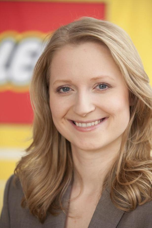 Katharina Sasse, stellvertretende Pressesprecherin im Brand-Relations-Team von Lego DACH. (c: lego) Eine lächelnde Frau mit blonden Haaren vor einem gelb-roten Hintergrund mit dem Lego-Logo.