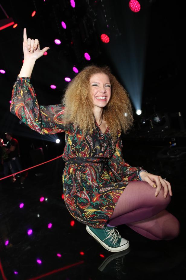 Eine Frau mit lockigem Haar und einem bunten Kleid kniet und lächelt.