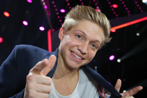 Ein lächelnder junger Mann mit blonden Haaren zeigt mit dem Finger auf die Kamera.