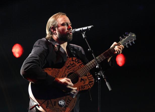 Ein Mann mit Bart spielt Gitarre und singt in ein Mikrofon. Die Gitarre ist mit Schriftzügen versehen.