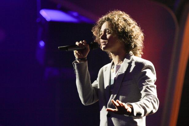 Ein Sänger mit lockigem Haar tritt auf einer Bühne mit Mikrofon auf.