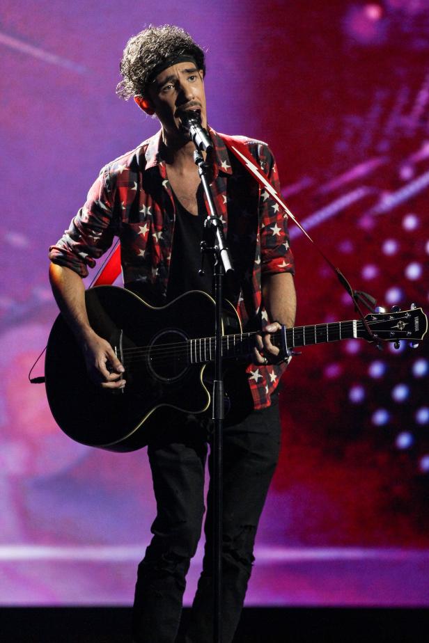 Ein Mann mit Stirnband singt und spielt Gitarre auf einer Bühne.