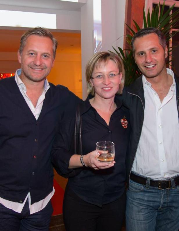 Drei Personen posieren für ein Foto, eine Frau hält ein Glas mit einer bernsteinfarbenen Flüssigkeit.