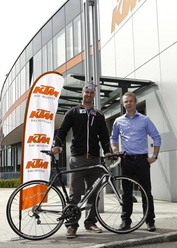 Zwei Männer posieren mit einem Rennrad vor einem Gebäude von KTM Bike Industries.