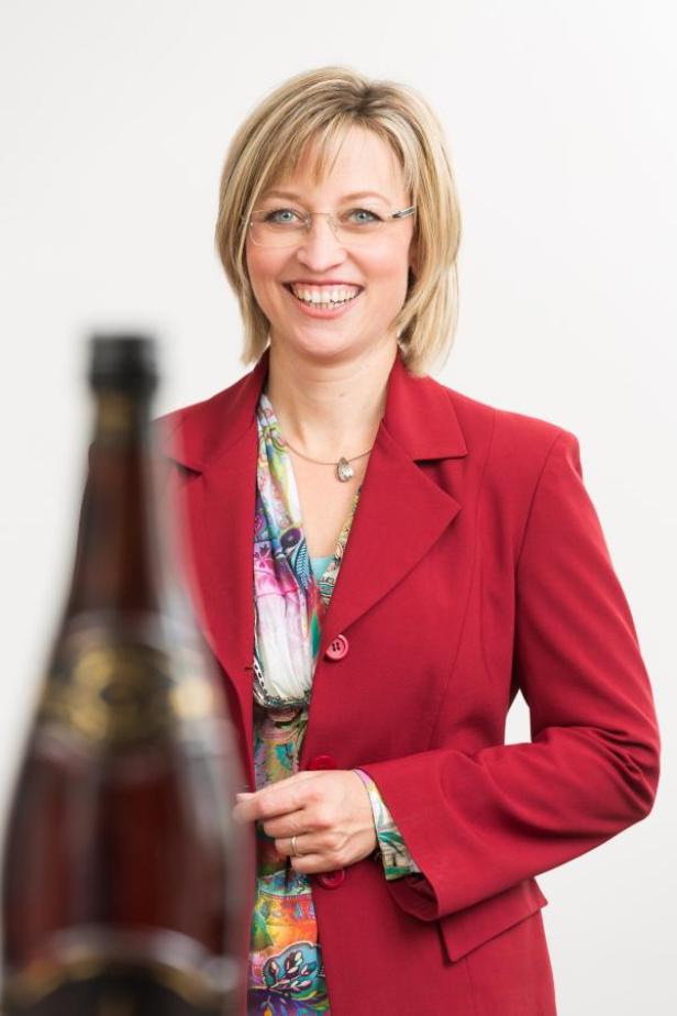 Eine lächelnde Frau mit Brille und rotem Blazer steht vor einer Champagnerflasche.