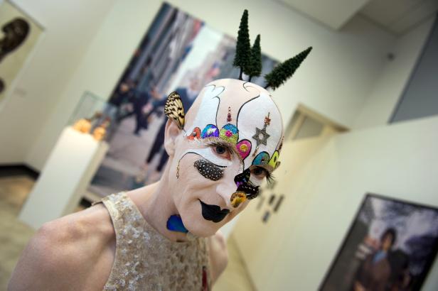 Eine Person mit aufwendigem Make-up und kleinen Bäumen auf dem Kopf in einer Galerie.