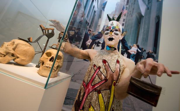 Ein Mann mit aufwendigem Make-up und Kostüm posiert vor einer Museumsausstellung mit Schädeln.