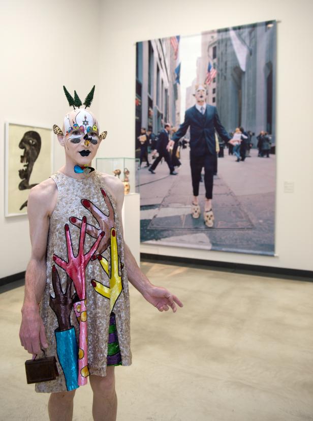 Ein Mann in einem verzierten Kleid posiert vor einem Kunstwerk in einer Galerie.