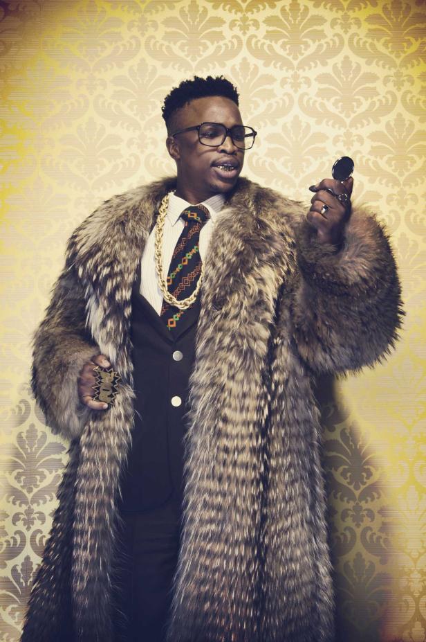 Ein Mann mit Goldzähnen, Pelzmantel und großer Goldkette posiert vor einer gemusterten Tapete.