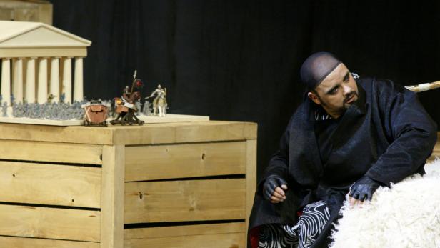 Ein Schauspieler in einem schwarzen Kimono sitzt neben einem Modell eines griechischen Tempels.