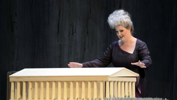 Eine Opernsängerin mit weiß gelockter Perücke steht vor einem Modell eines griechischen Tempels.