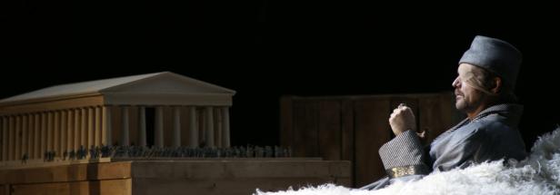 Ein blinder Mann in einem Kostüm sitzt vor einem Modell eines griechischen Tempels.