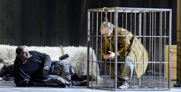 Ein Mann sitzt in einem Käfig, während ein anderer daneben auf dem Boden liegt.