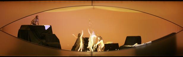 Auf einer Bühne stehen drei Frauen in goldenen Kostümen, während Konfetti auf sie herabregnet. Ein Mann steht abseits auf einem Podest.