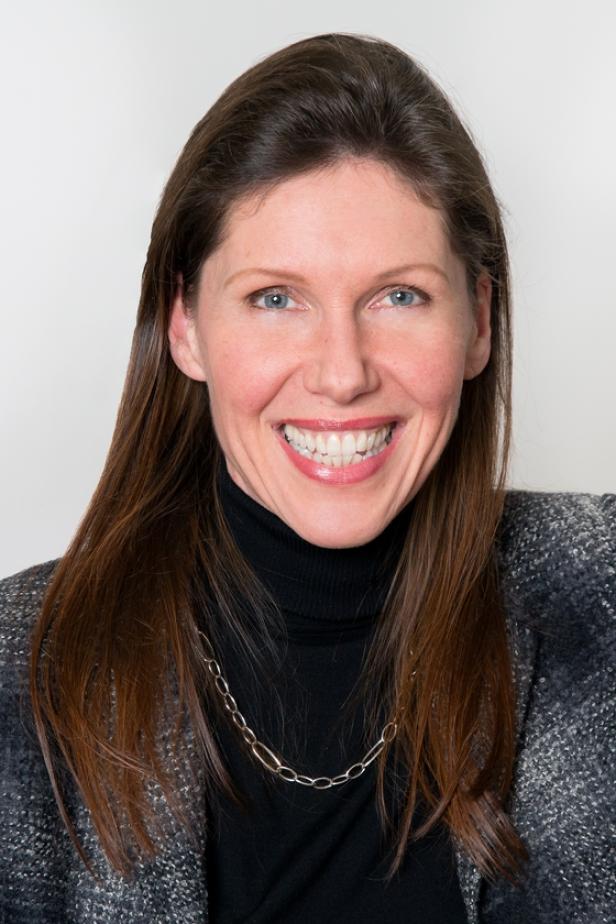 Porträt einer lächelnden Frau mit braunen Haaren, die einen schwarzen Rollkragenpullover und eine graue Jacke trägt.