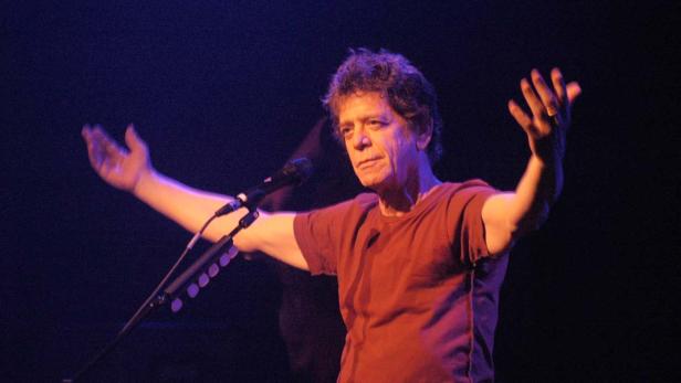 Lou Reed steht mit erhobenen Armen auf einer Bühne.