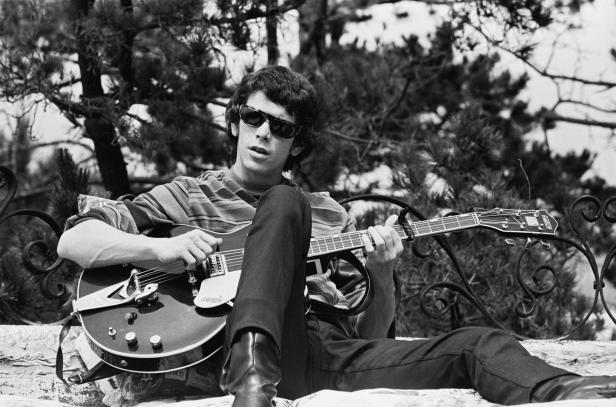 Lou Reed sitzt mit Sonnenbrille und Gitarre im Freien.