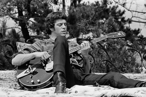 Lou Reed sitzt entspannt im Freien und spielt Gitarre.