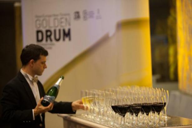 Ein Kellner schenkt Sekt in ein Glas vor dem Hintergrund des „Golden Drum“-Logos ein.