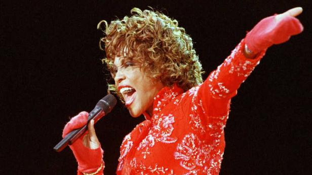 Whitney Houston singt in einem roten Kleid mit erhobenem Arm.
