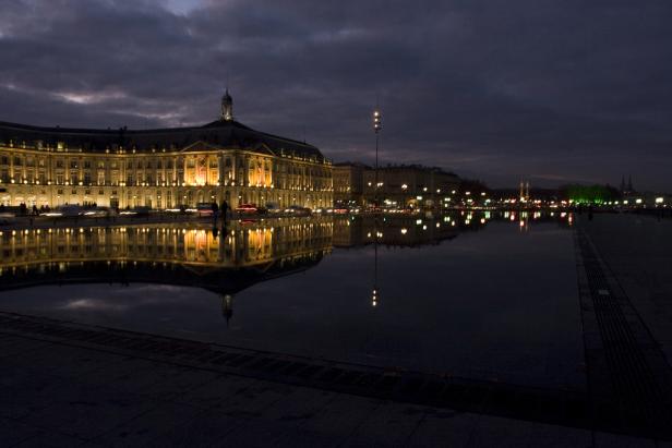 Das beleuchtete Gebäude der Place de la Bourse spiegelt sich im Wasser bei Nacht.