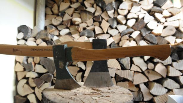 Zwei Äxte stehen auf einem Holzscheit vor einem Stapel Brennholz.
