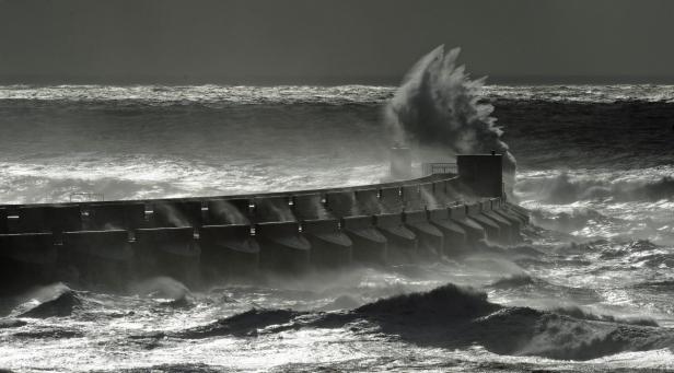 Eine hohe Welle bricht an einem Pier und spritzt hoch in die Luft.