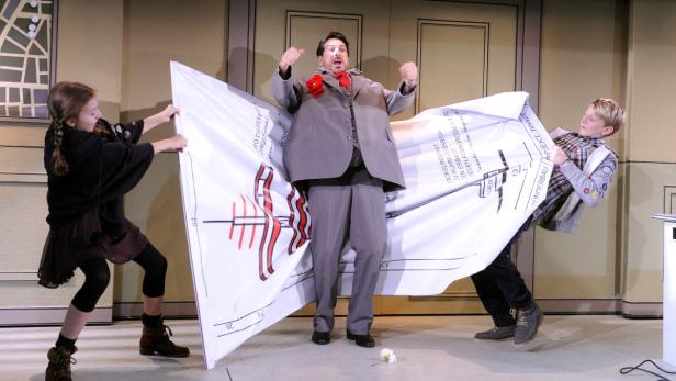 Ein Mann im Anzug steht auf einer Bühne, während zwei Kinder ein großes Banner mit einer technischen Zeichnung halten.