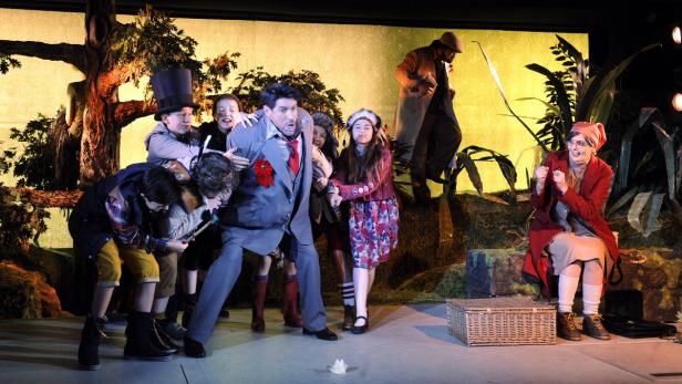 Eine Theaterszene mit einer Gruppe von Kindern, die einen Mann in einem Anzug umringen.