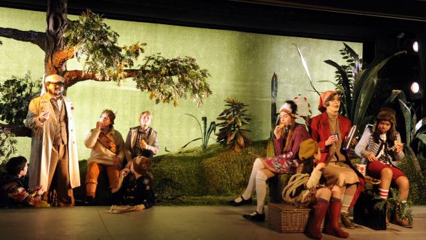 Eine Gruppe von Kindern und ein Mann in Kostümen sitzen vor einer Bühnenkulisse.