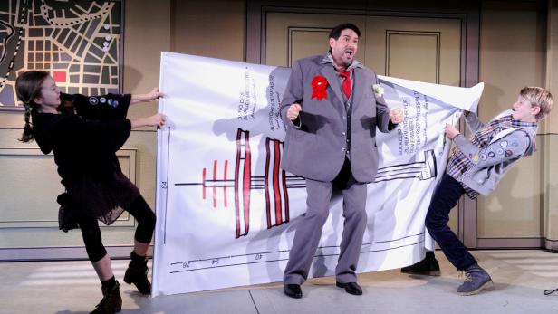 Ein Mann in einem übergroßen Anzug steht zwischen zwei Kindern, die eine Bauzeichnung halten.