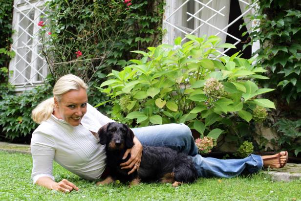 Eine Frau liegt mit einem Dackel im Gras vor einem mit Efeu bewachsenen Haus.