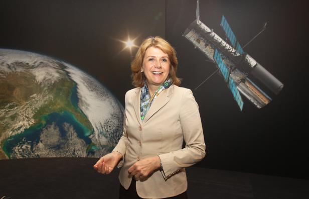 Eine Frau steht vor einem Hintergrund mit Erde und dem Hubble-Weltraumteleskop.