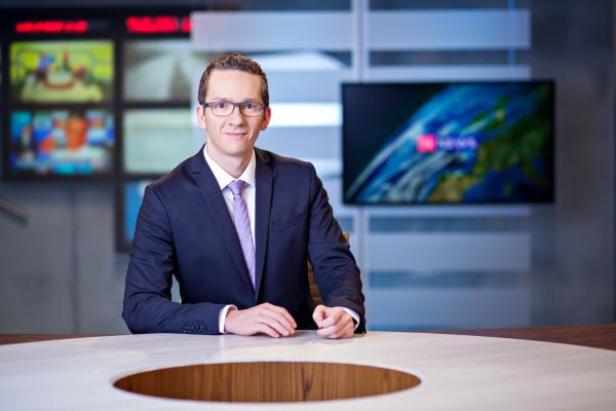 Ein Mann mit Brille und Anzug sitzt an einem Tisch in einem Fernsehstudio.