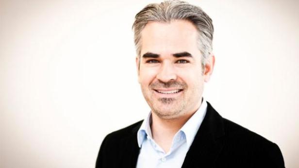 Ein Mann mit grau meliertem Haar lächelt in einem schwarzen Sakko.