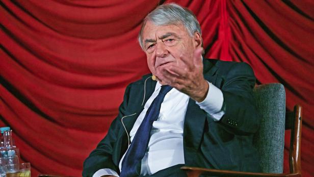 Ein Mann in einem Anzug gestikuliert vor einem roten Vorhang.