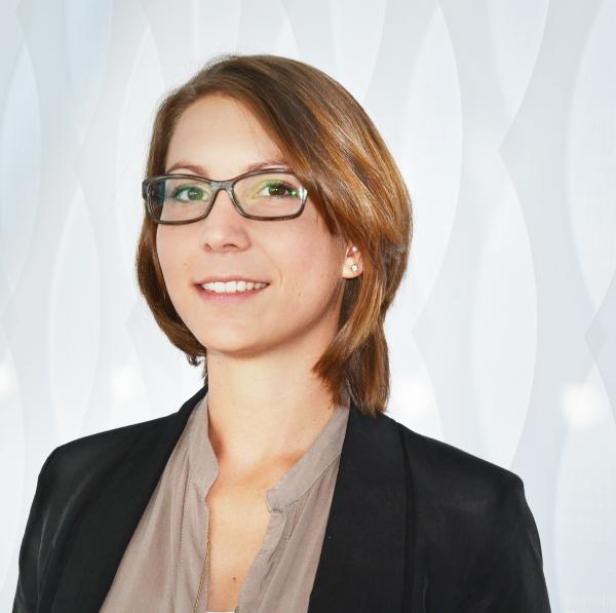 Eine lächelnde Frau mit Brille und braunem Haar vor einem hellen Hintergrund.