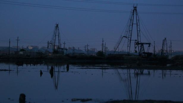 Mehrere Ölfördertürme spiegeln sich im Wasser in der Abenddämmerung.
