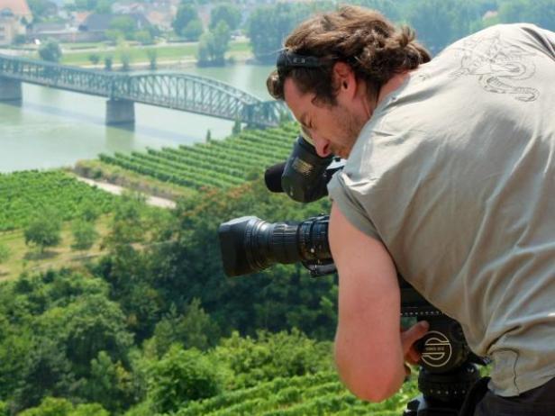 Ein Kameramann filmt eine Landschaft mit Weinbergen und einer Brücke.