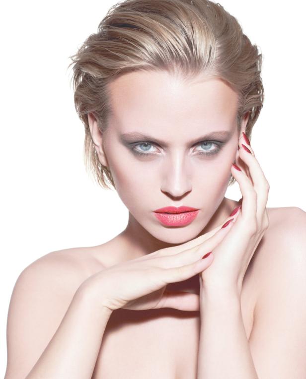 Eine Frau mit blonden, nach hinten gekämmten Haaren und auffälligem Make-up posiert vor einem weißen Hintergrund.