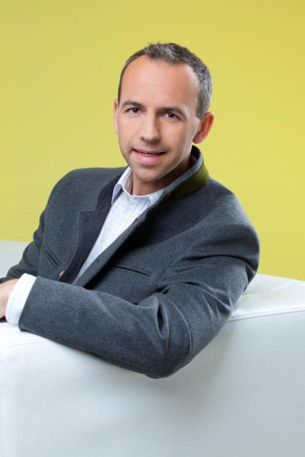 Ein Mann in Trachtenjacke lehnt auf einem weißen Sofa vor gelbem Hintergrund.