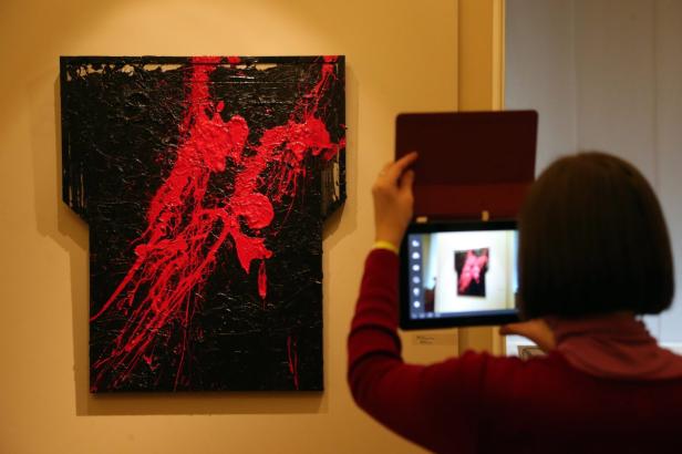 Eine Person fotografiert ein abstraktes Gemälde mit roter Farbe auf schwarzem Grund.