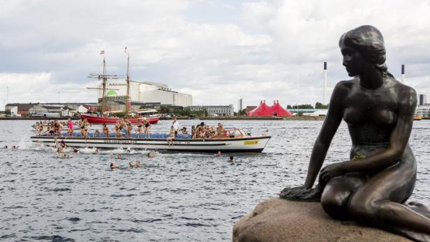 Die kleine Meerjungfrau sitzt auf einem Felsen, während im Hintergrund Menschen von einem Boot ins Wasser springen.