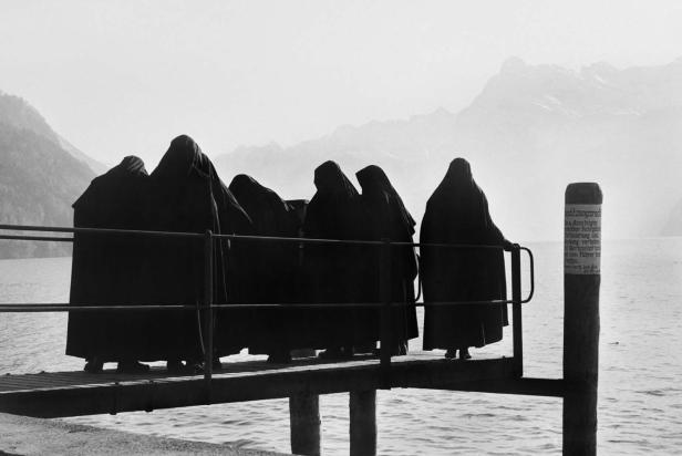 Eine Gruppe Nonnen steht auf einem Steg an einem See vor einer Bergkulisse.