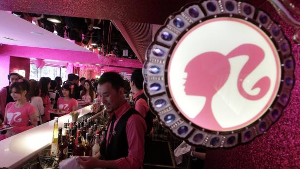 In einem Barbie-Themenrestaurant bedient ein Barkeeper Gäste vor einem Barbie-Logo.