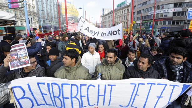Eine Demonstration mit Menschen, die Schilder mit der Aufschrift „Asyl in Not“ und „Bleiberecht Jetzt“ tragen.