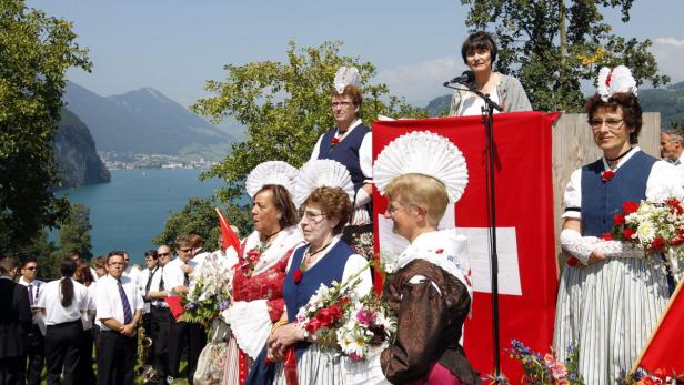 Frauen in Tracht und eine Rednerin vor einer Schweizer Flagge mit Bergpanorama.