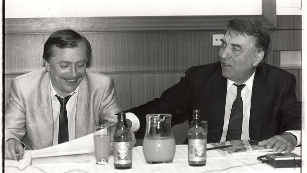 Zwei Männer sitzen an einem Tisch mit Getränken und Dokumenten.